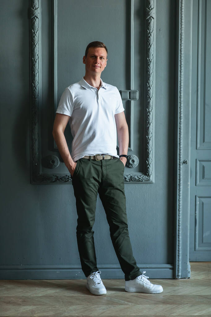 Guapo hombre alto con estilo en un polo, pantalones verdes y zapatillas blancas de pie por la pared en una habitación con interior clásico y mirando a la cámara complacido - Foto, Imagen