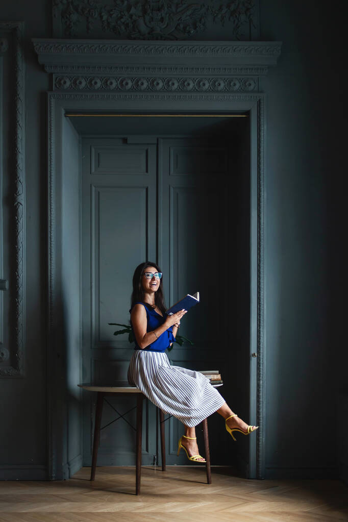 Mulher de cabelos compridos bonita elegante uma camisa azul e saia sentada em óculos na mesa com um livro em mãos no interior do compartimento, olhando para a câmera e rindo
 - Foto, Imagem