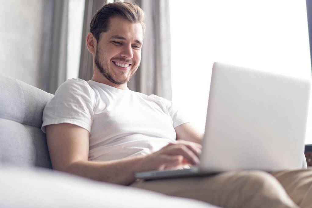 Neue Blogpost eintippen. Schöner junger Mann benutzt seinen Laptop mit einem Lächeln, während er zu Hause auf der Couch sitzt. - Foto, Bild