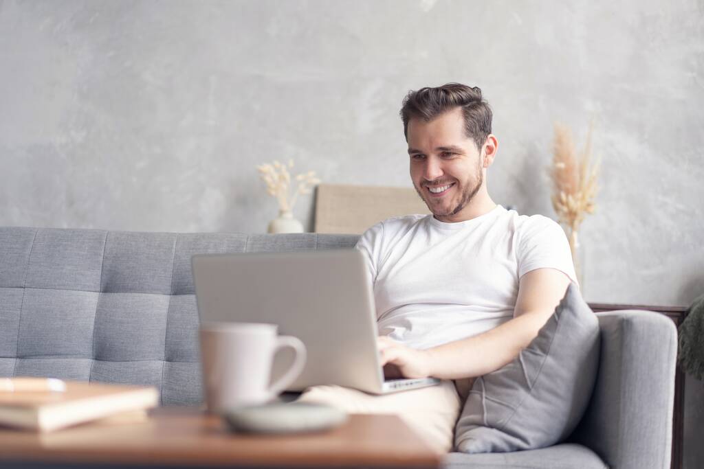 Yeni blog yazıları yazılıyor. Yakışıklı genç adam evdeki kanepede otururken bilgisayarını gülümseyerek kullanıyor.. - Fotoğraf, Görsel