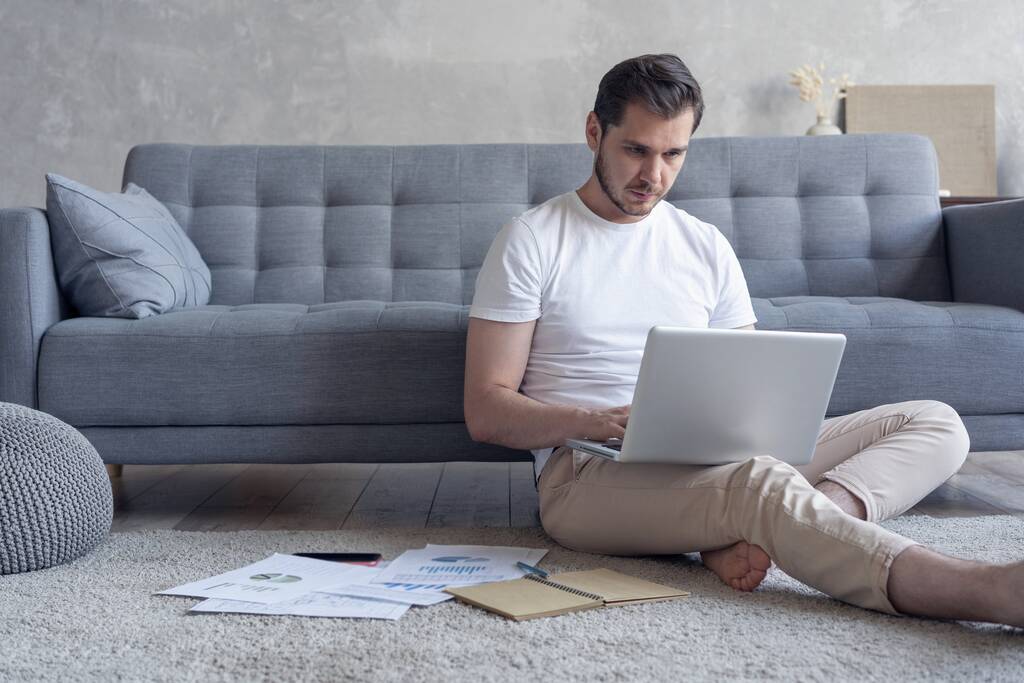 Man werkt op laptop computer zittend op de vloer met zijn dagboek en officiële papieren thuis - Foto, afbeelding