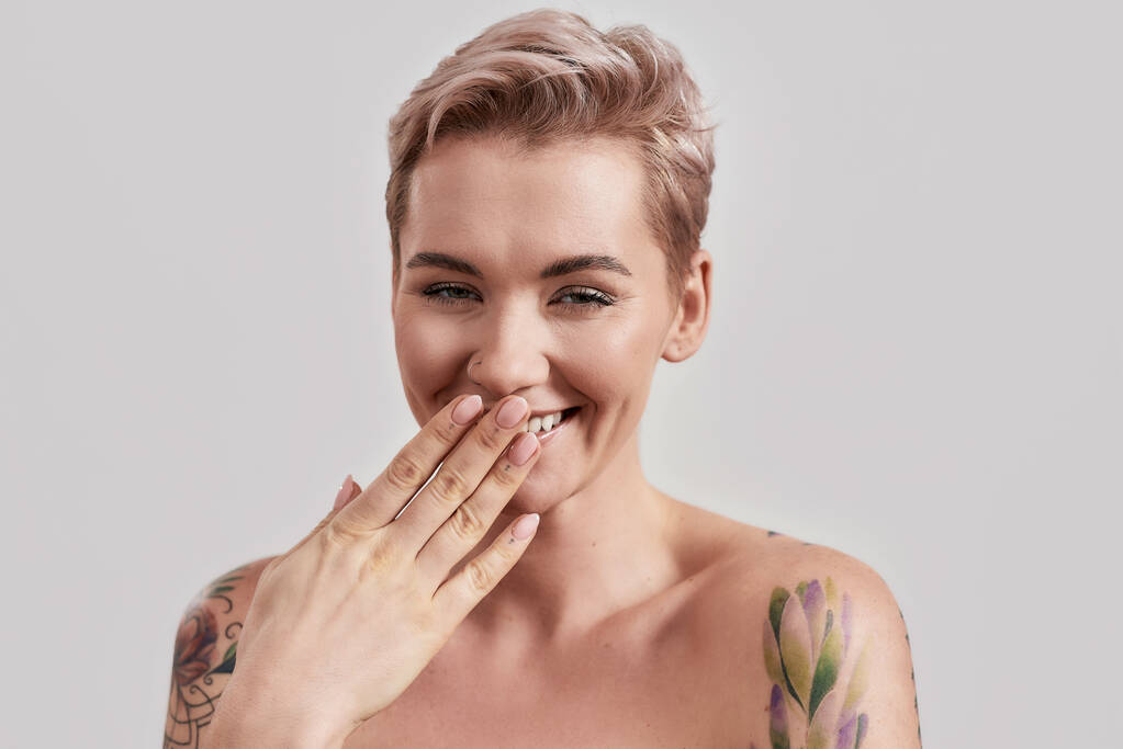 Qué curioso. Retrato de una joven atractiva mujer semidesnuda tatuada con la piel perfecta riéndose de la cámara, cubriendo la boca con la mano aislada sobre un fondo claro - Foto, Imagen