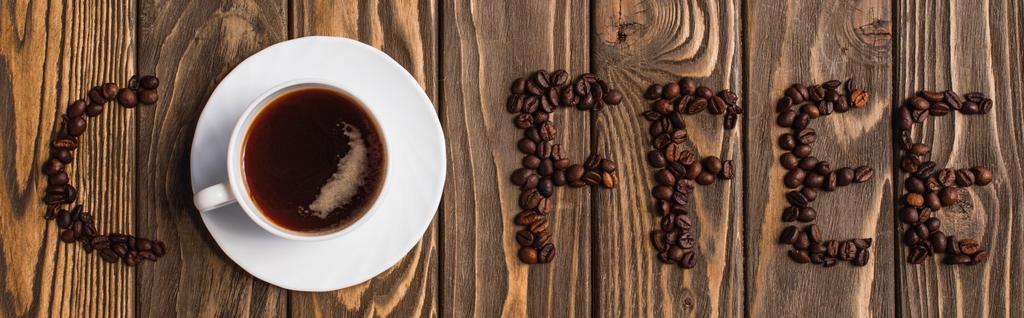top view of cup of coffee on saucer and coffee lettering made of beans on wooden surface, panoramic shot - Photo, Image