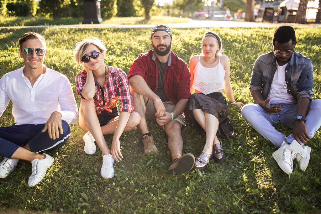 Różnorodna grupa wesołych przyjaciół siedzi na trawie o zachodzie słońca w parku przyrody. - Zdjęcie, obraz