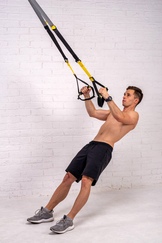Hombre musculoso haciendo ejercicios con correas trx, cerca de una pared de ladrillo blanco. Culturismo. - Foto, imagen