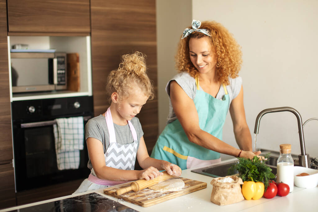 Красивая молодая мама и ее милая маленькая дочь прокатывают тесто дома. - Фото, изображение