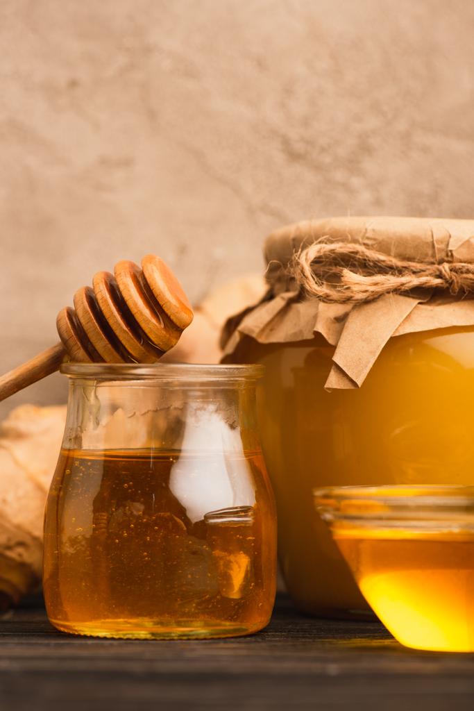 close up view of sweet honey in glass jars with stick on wooden surface near beige concrete wall - Photo, Image