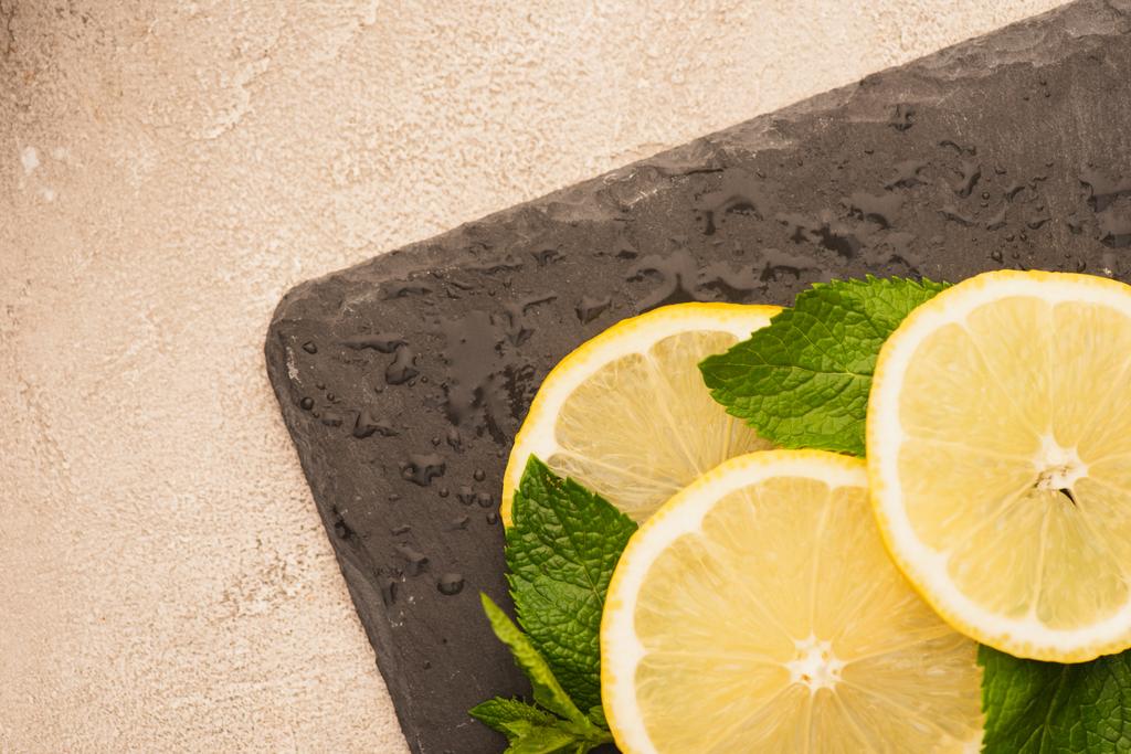 vista superior de hojas verdes menta, rodajas de limón sobre pizarra negra sobre superficie de hormigón beige - Foto, imagen