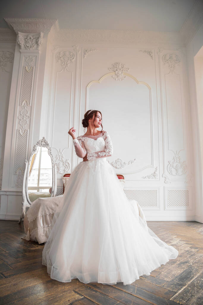 .Ολόσωμο πορτραίτο νύφης σε φωτεινό στούντιο με φως από το παράθυρο. Το κορίτσι στέκεται επιθετικά κοιτάζοντας προς τα πλάγια. Μακιγιάζ γάμου και styling - Φωτογραφία, εικόνα
