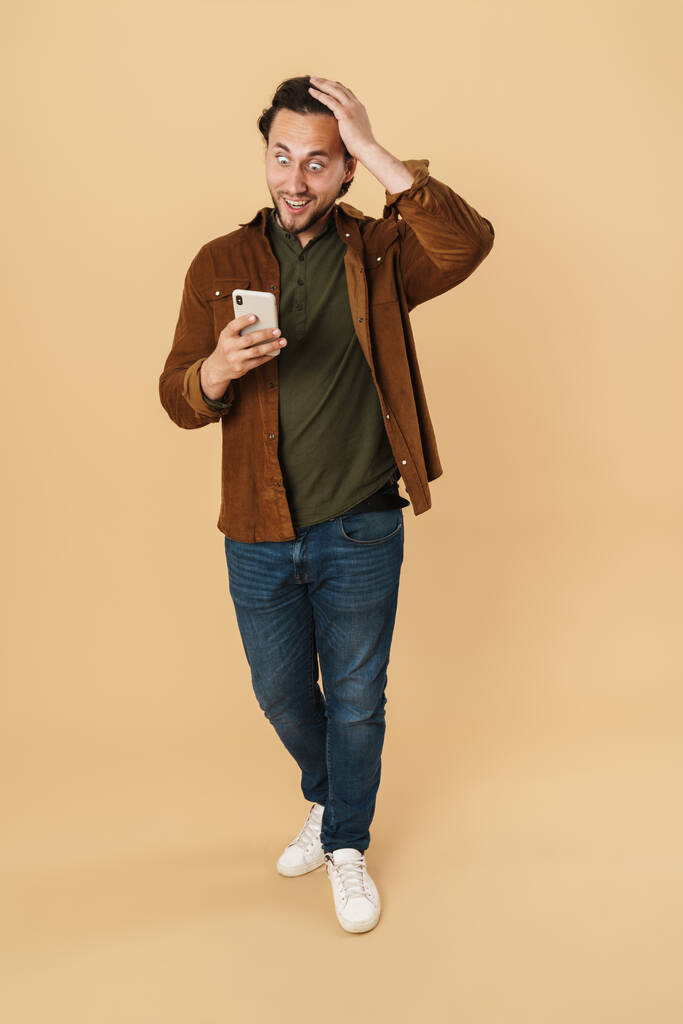Bild eines erstaunten Mannes, der seine Überraschung zum Ausdruck bringt, während er sein Handy isoliert über beigem Hintergrund benutzt - Foto, Bild
