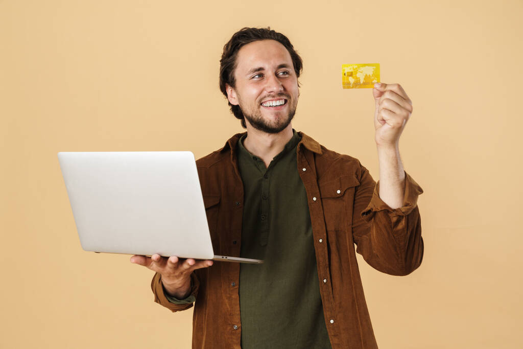 Kép elégedett borotválatlan férfi kezében hitelkártya használata közben laptop izolált bézs háttér - Fotó, kép