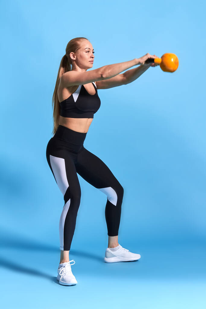 молодая привлекательная спортсменка делает упражнения колокольчик чайник на синем фоне. - Фото, изображение