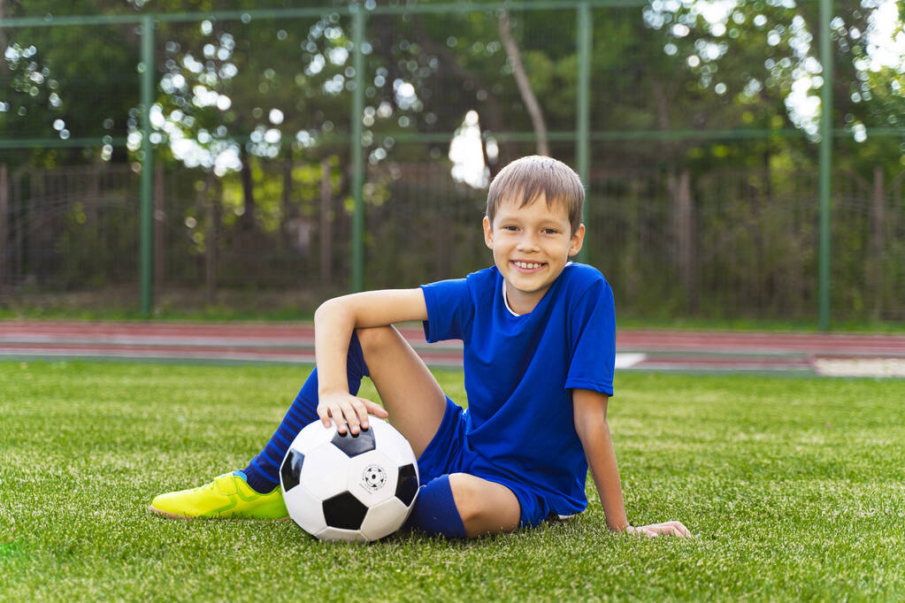 Ένα μικρό αγόρι κάθεται σε ένα πράσινο γήπεδο ποδοσφαίρου, μια μπάλα ποδοσφαίρου. - Φωτογραφία, εικόνα