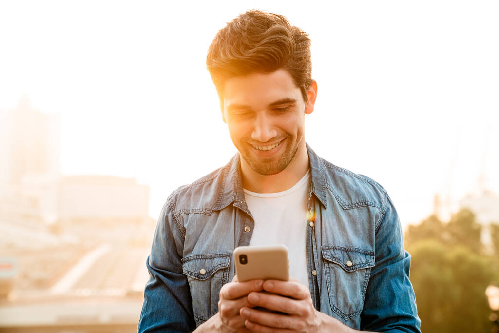 Foto de hombre guapo alegre sonriendo y usando el teléfono móvil mientras camina al aire libre - Foto, Imagen
