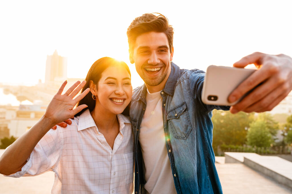 Imagen de pareja multicultural feliz saludando de la mano y tomando foto selfie en el teléfono celular al aire libre - Foto, Imagen