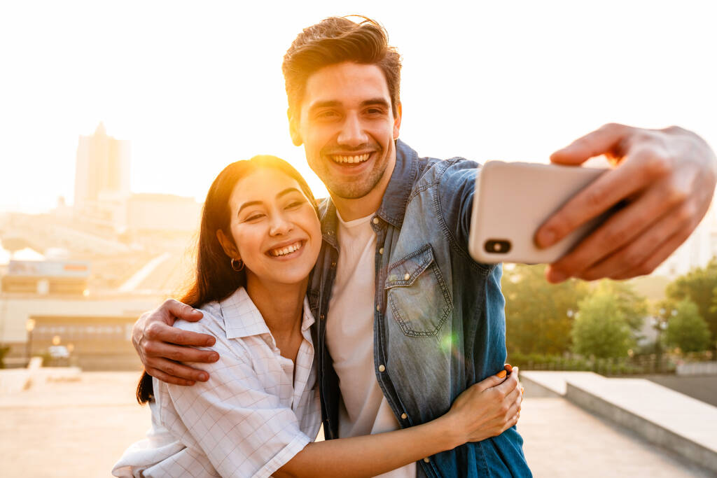 Imagem de casal multicultural sorridente abraçando e tirando foto selfie no celular ao ar livre
 - Foto, Imagem