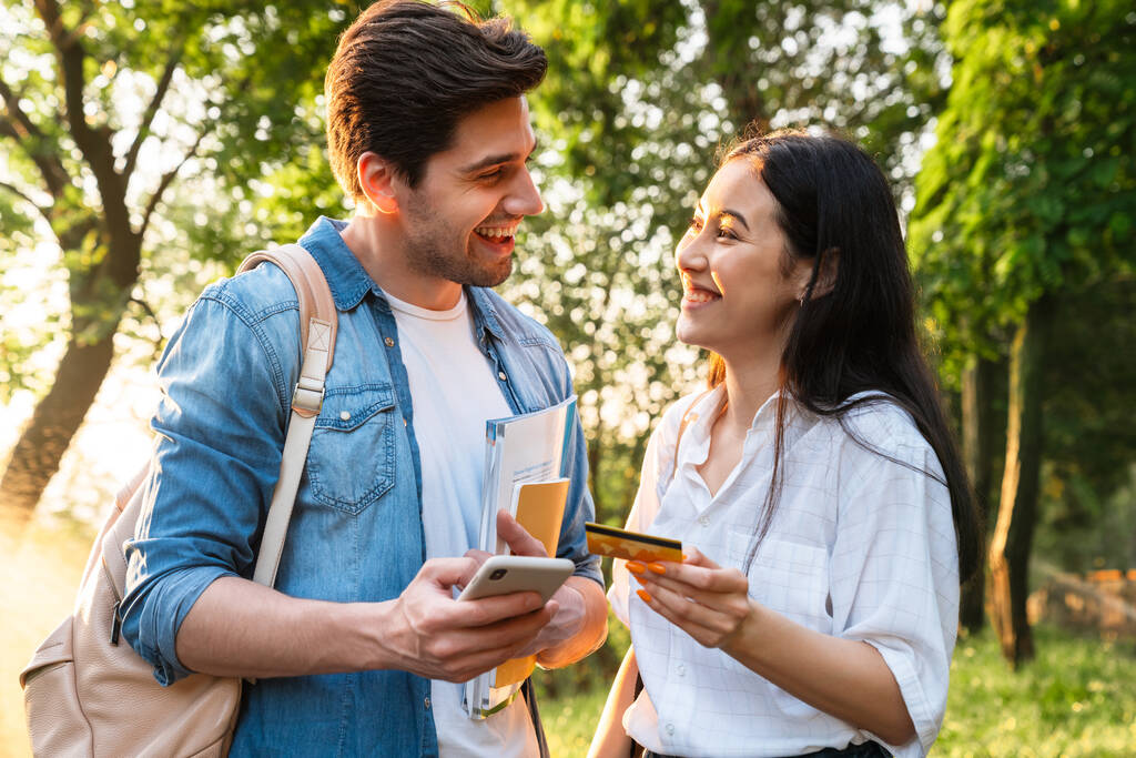 Imagem de casal estudantil multicultural alegre usando telefone celular e segurando cartão de crédito enquanto caminhava no parque verde
 - Foto, Imagem