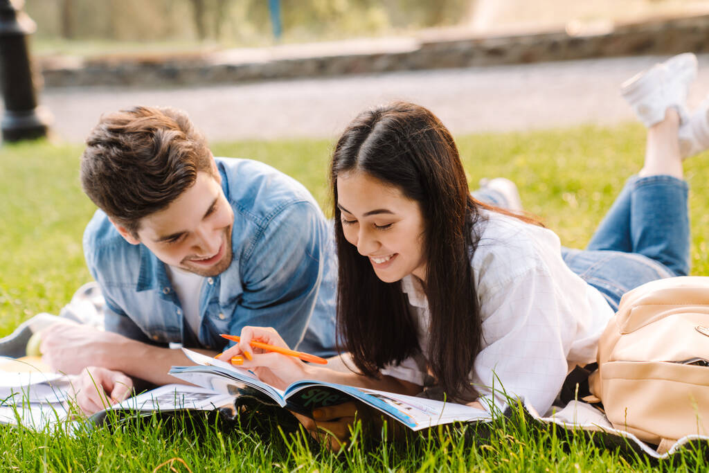 Immagine di gioiosa coppia di studenti multiculturali che fanno i compiti e sorridono sdraiati sull'erba nel parco - Foto, immagini