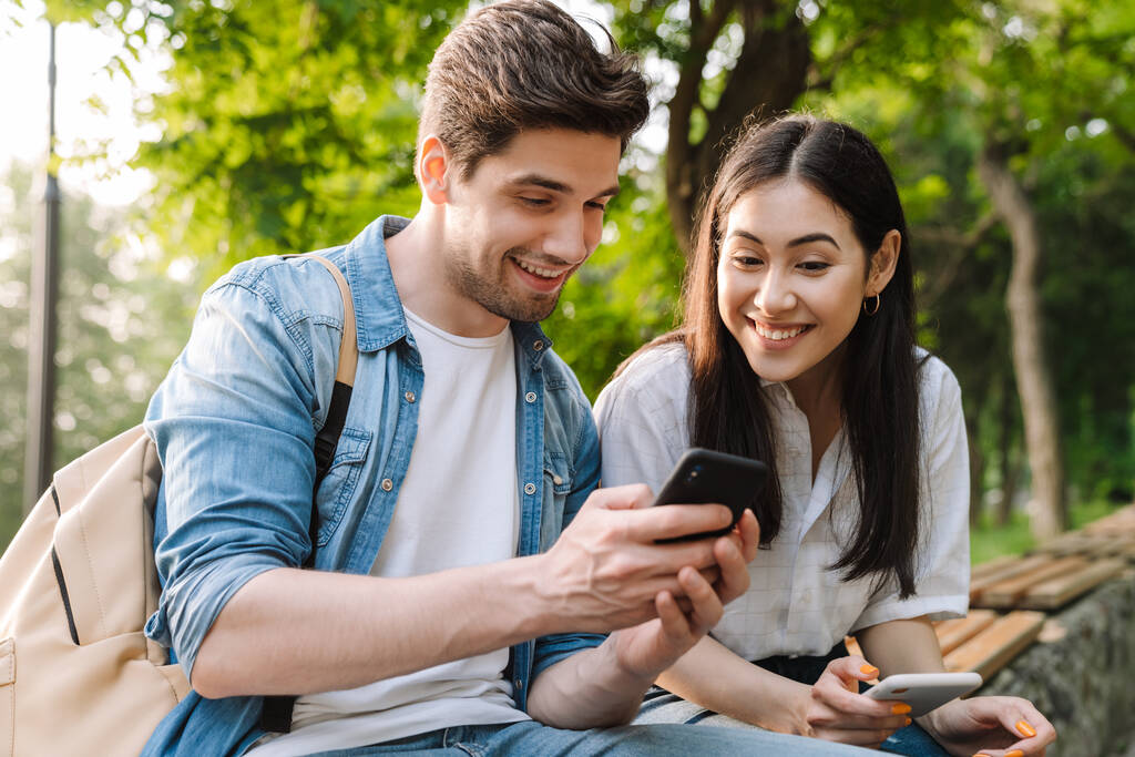 Imagem de casal multicultural satisfeito usando telefones celulares e sorrindo enquanto sentado no banco no parque
 - Foto, Imagem