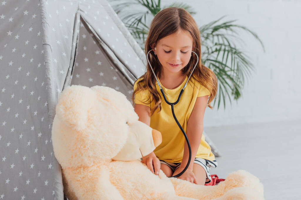 girl in yellow t-shirt playing doctor with stethoscope and teddy bear near play tent - Photo, Image