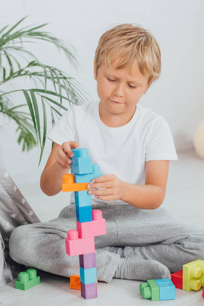 niño concentrado en pijamas sentado en el suelo y jugando con bloques de construcción de colores - Foto, Imagen