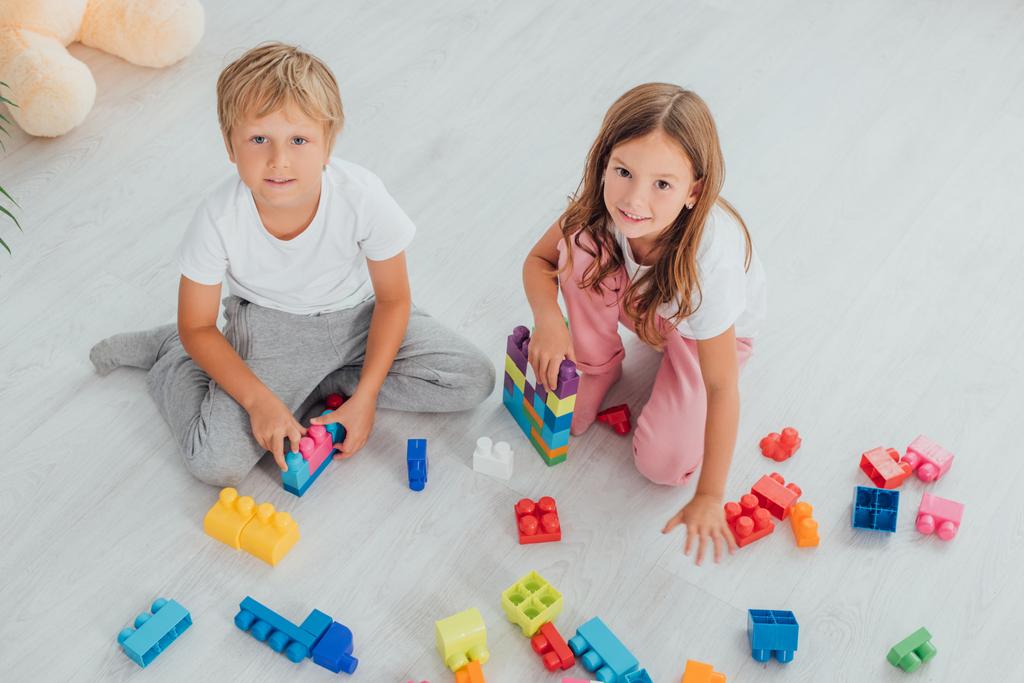 high angle view of children in pajamas sitting on floor near building blocks and looking at camera - Photo, Image