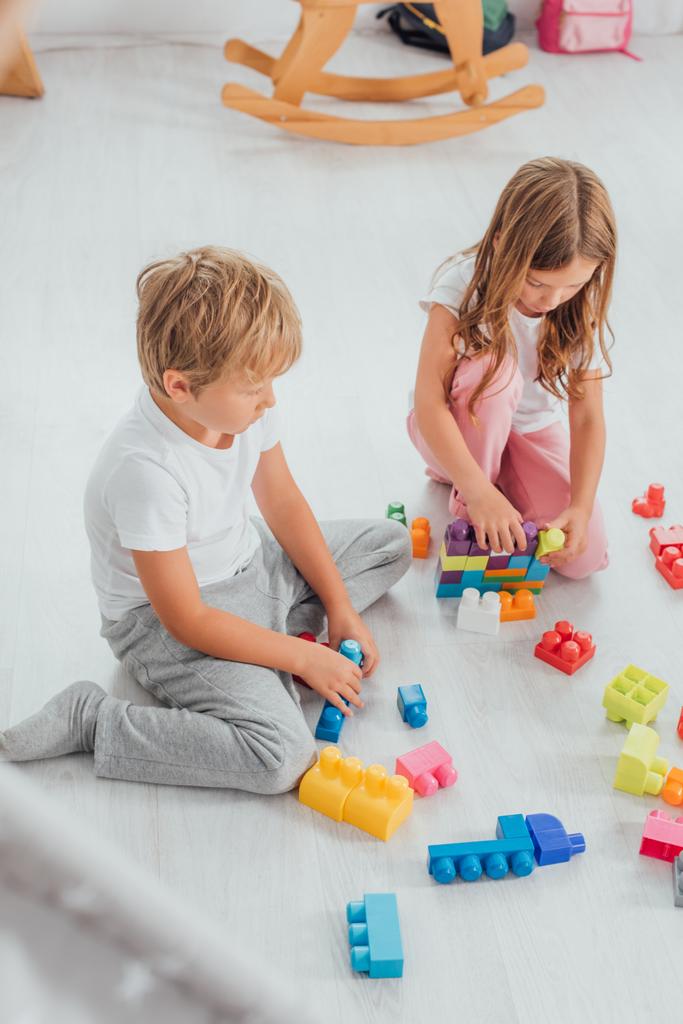 vysoký úhel pohledu na koncentrované sourozence v pyžamu sedící na podlaze a hrající si s stavebními kameny - Fotografie, Obrázek