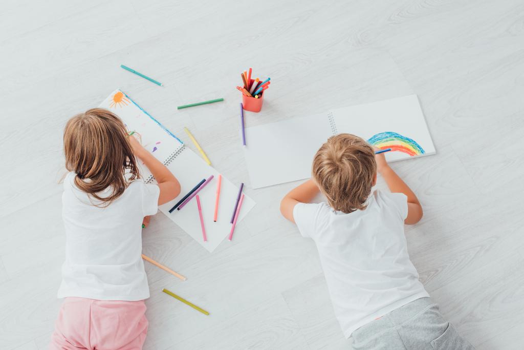 パジャマ姿の兄と妹が床に横になってスケッチブックスのペンで絵を描く - 写真・画像