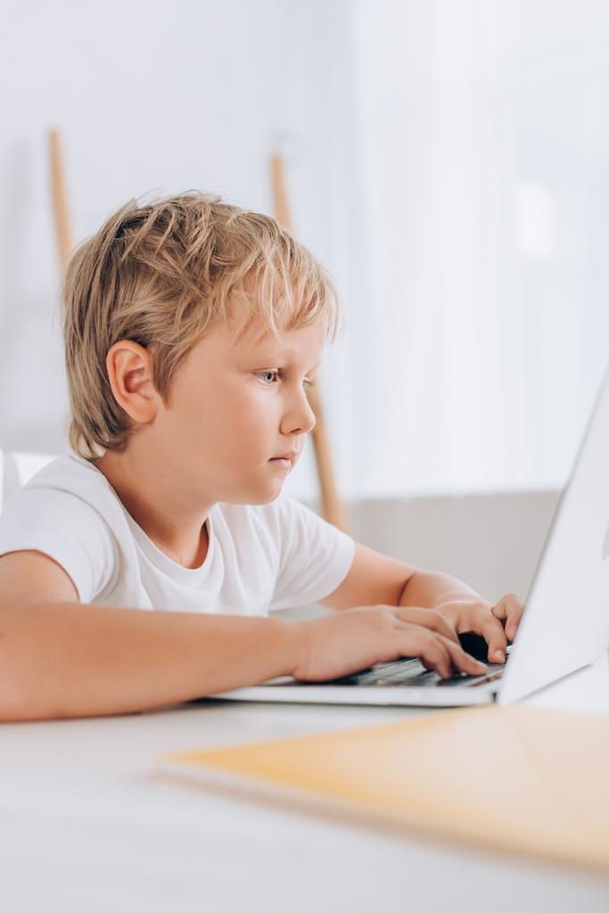 selektivní zaměření zaměřeného dítěte v bílém tričku pomocí notebooku při sezení u stolu - Fotografie, Obrázek