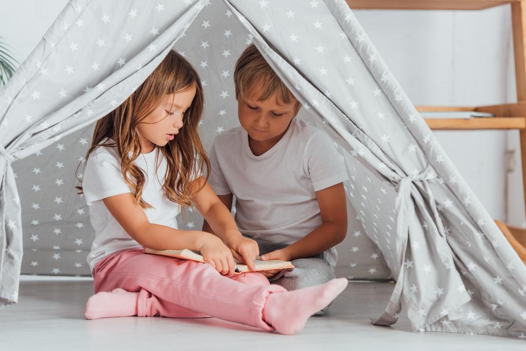 brother and sister in pajamas sitting on floor in kids wigwam and reading book - Photo, Image