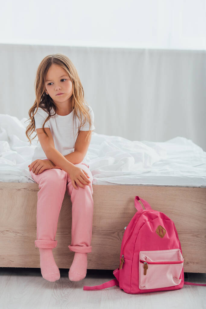 upset girl in pajamas sitting on bed near school backpack on floor - Photo, Image