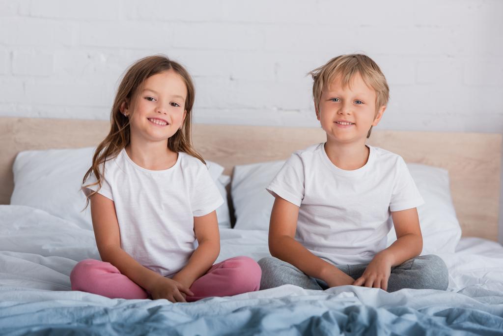 Bruder und Schwester im Schlafanzug schauen in die Kamera, während sie mit gekreuzten Beinen auf dem Bett sitzen - Foto, Bild