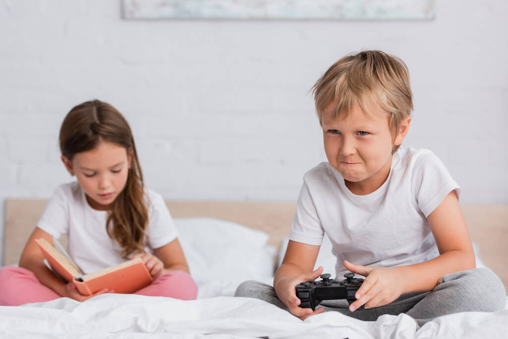 KYIV, UCRANIA - 21 de julio de 2020: enfoque selectivo del niño concentrado jugando videojuegos cerca del libro de lectura hermana en el dormitorio - Foto, Imagen