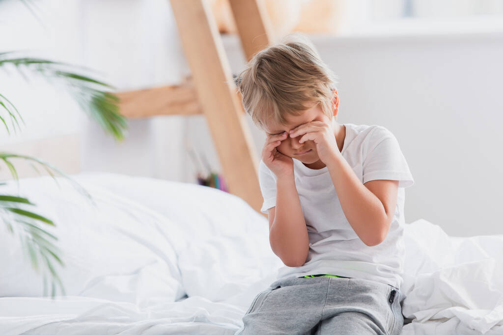 Проснувшийся мальчик касается глаз, сидя на кровати в пижаме - Фото, изображение