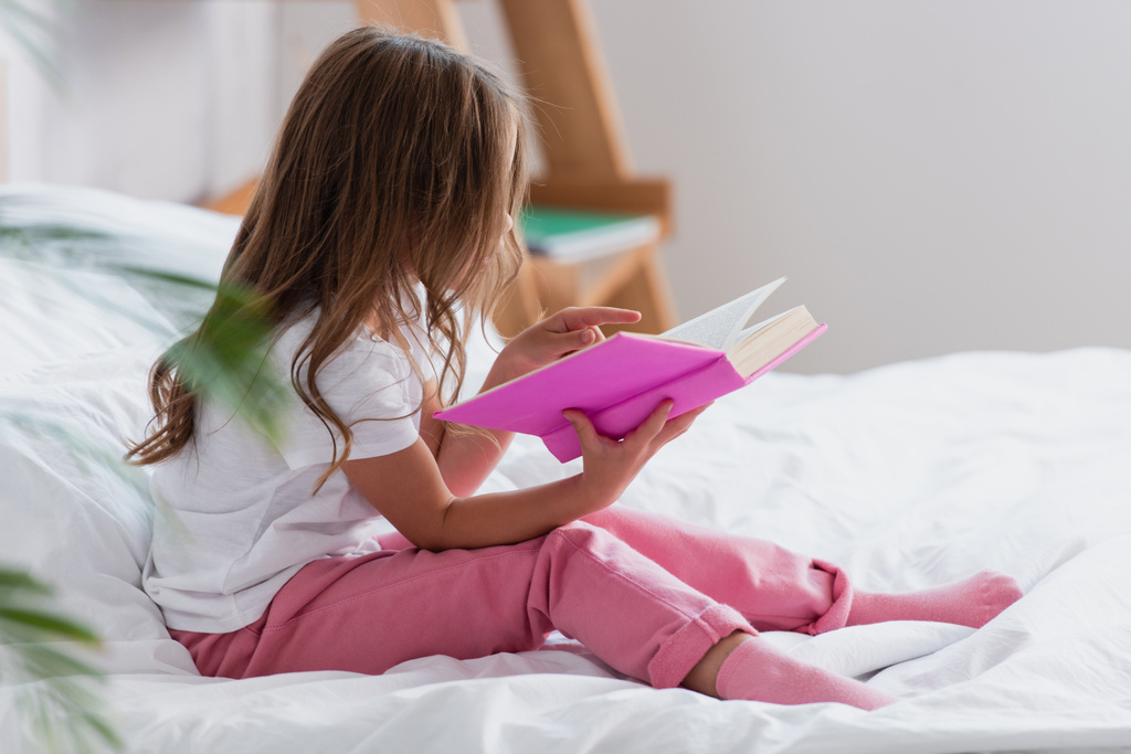 Pijamalı bir kız. Yatakta oturuyor ve sabah kitap okuyor. - Fotoğraf, Görsel