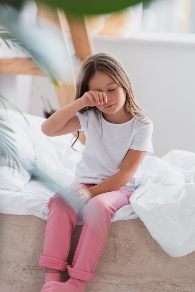 επιλεκτική εστίαση της αφύπνισης κορίτσι με πιτζάμες συγκινητικό μάτι, ενώ κάθεται στο κρεβάτι - Φωτογραφία, εικόνα