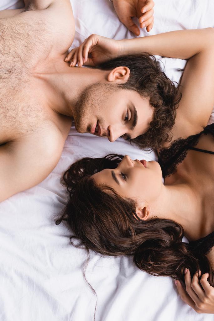 vista superior de hombre y mujer sin camisa en sujetador mirándose en la cama en casa - Foto, Imagen