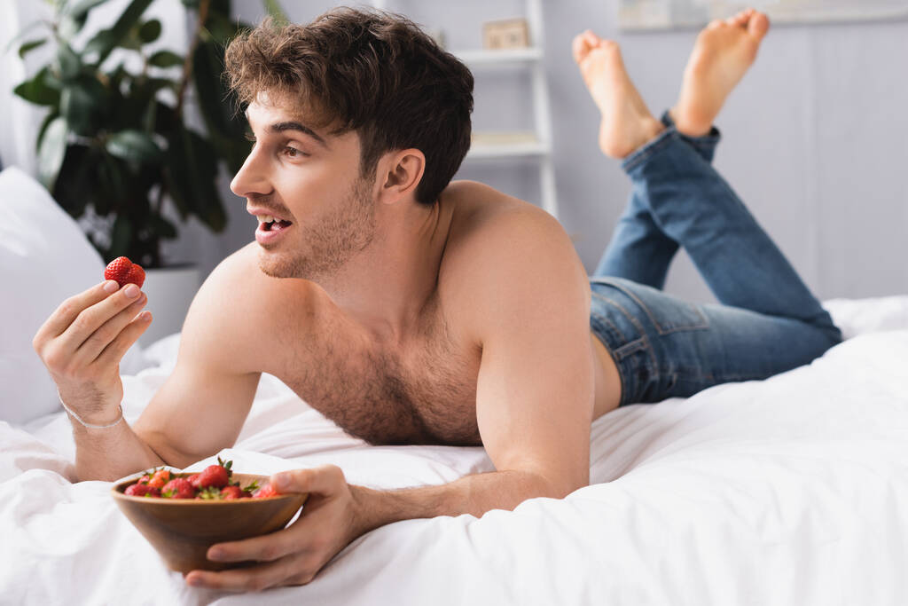 foco seletivo do homem sem camisa e descalço deitado na cama e segurando tigela com morangos maduros
  - Foto, Imagem