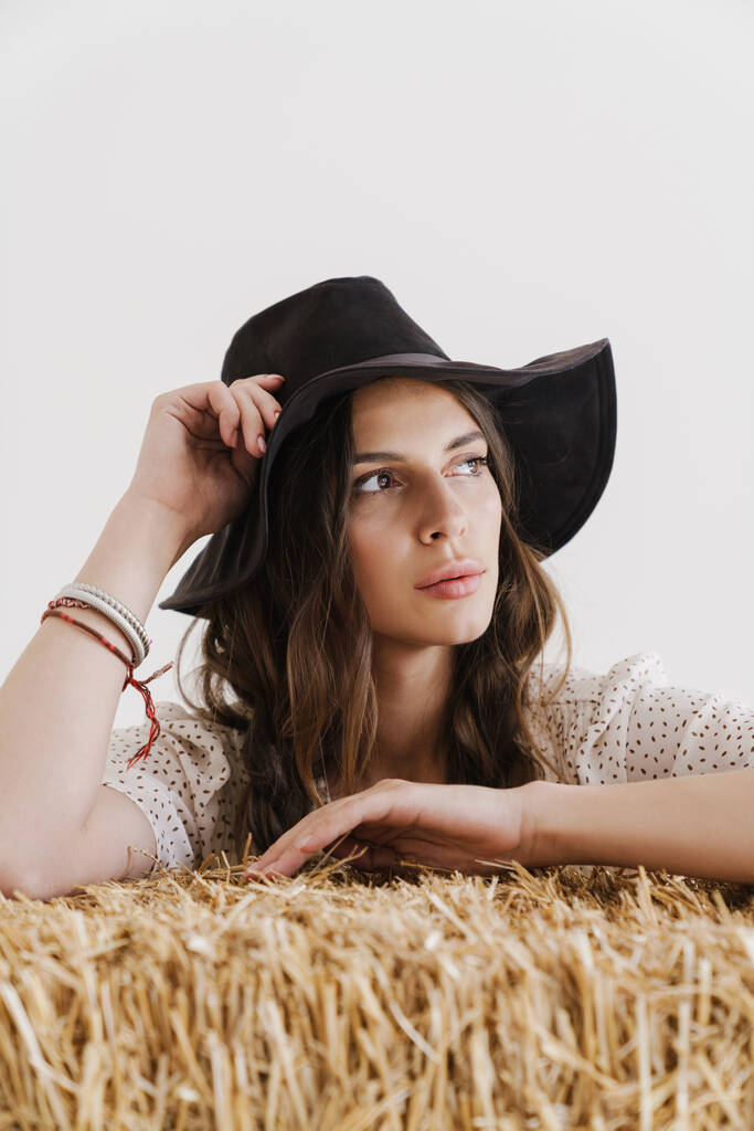 Фото молодой соблазнительной женщины в шляпе, смотрящей в сторону, опираясь на сено, изолированное на белом фоне - Фото, изображение
