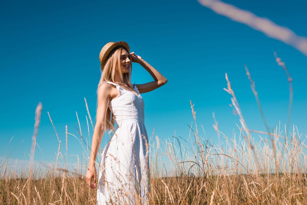 selective focus of stylish young woman in white dress touching straw hat and looking away against blue sky - Photo, Image