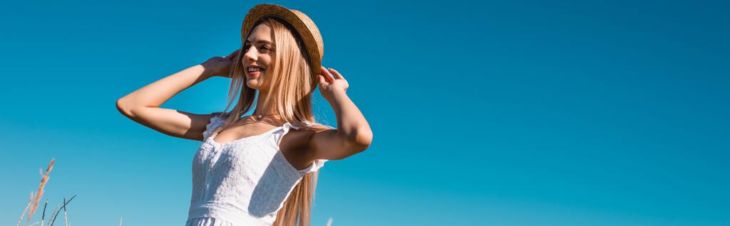 matala kulma näkymä nuori blondi nainen koskettaa olki hattu katsellen pois sinistä taivasta vasten, vaakasuora kuva - Valokuva, kuva
