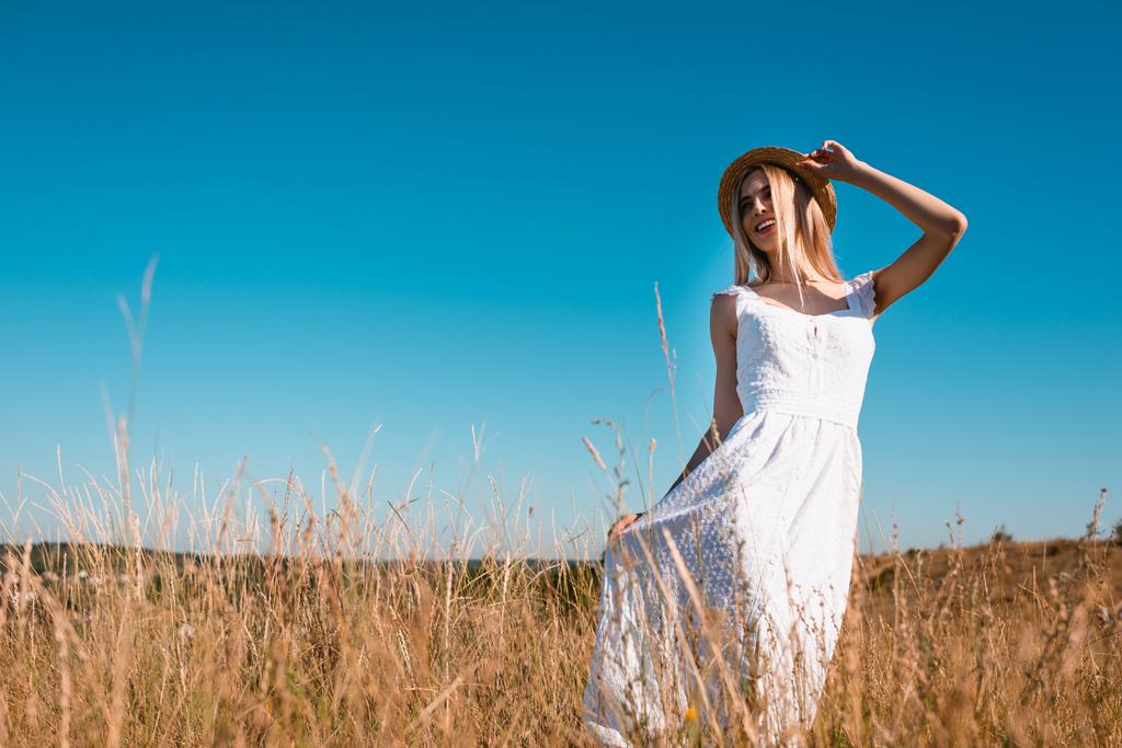 foco seletivo de mulher loira elegante tocando chapéu de palha e vestido branco enquanto posando no prado gramado contra o céu azul - Foto, Imagem