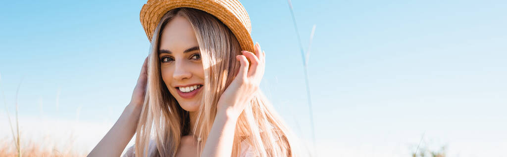 horizontaal beeld van jonge blonde vrouw aanraken strohoed terwijl kijken naar camera tegen blauwe hemel - Foto, afbeelding