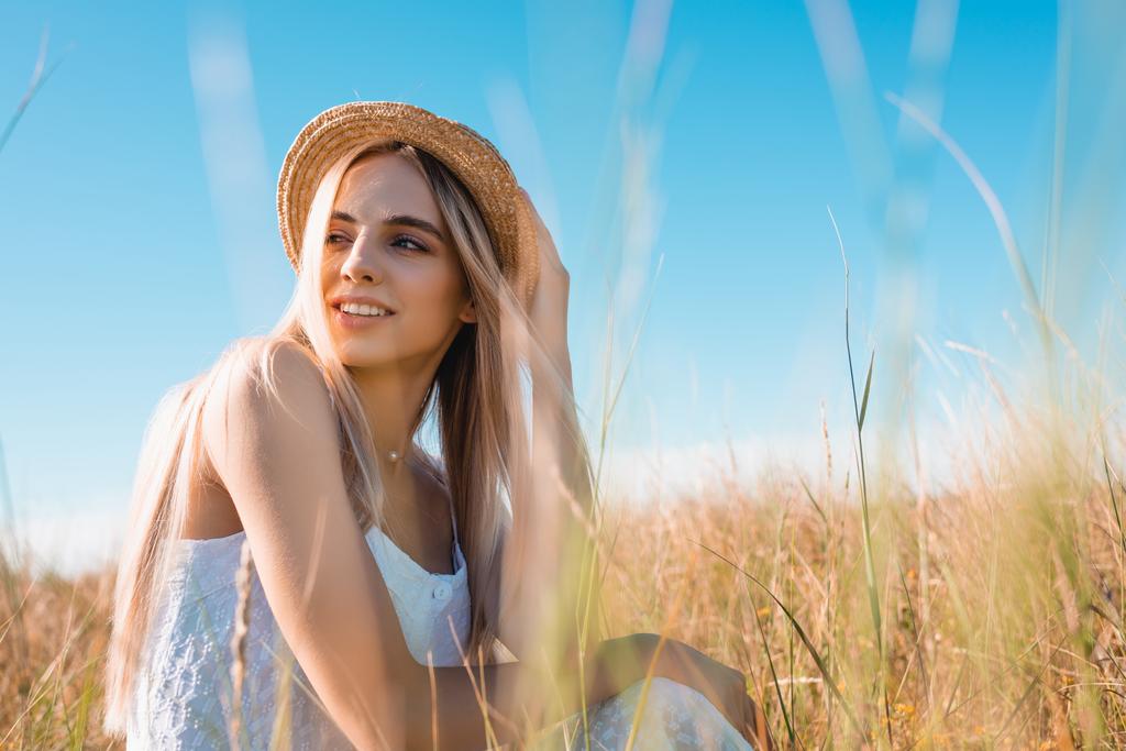 foyer sélectif de femme blonde élégante regardant loin tout en étant assis dans le champ et touchant chapeau de paille - Photo, image
