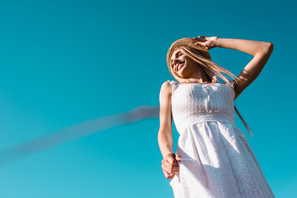 низкий угол зрения блондинка в белом платье касаясь соломенной шляпы против голубого неба - Фото, изображение