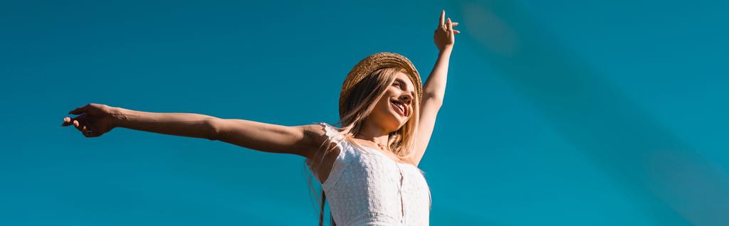horizontales Bild einer jungen Frau mit Strohhut, die mit ausgestreckten Händen vor blauem Himmel steht, Blick in den niedrigen Winkel - Foto, Bild