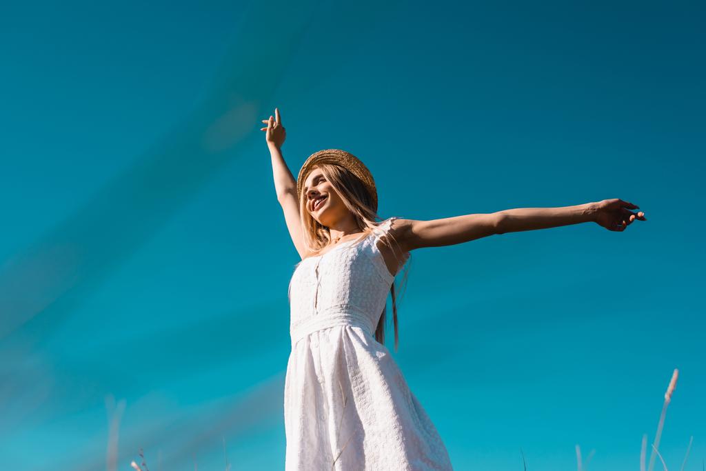 низький кут зору жінки в білій сукні і солом'яному капелюсі, дивлячись далеко, стоячи на блакитному небі з розтягнутими руками
 - Фото, зображення