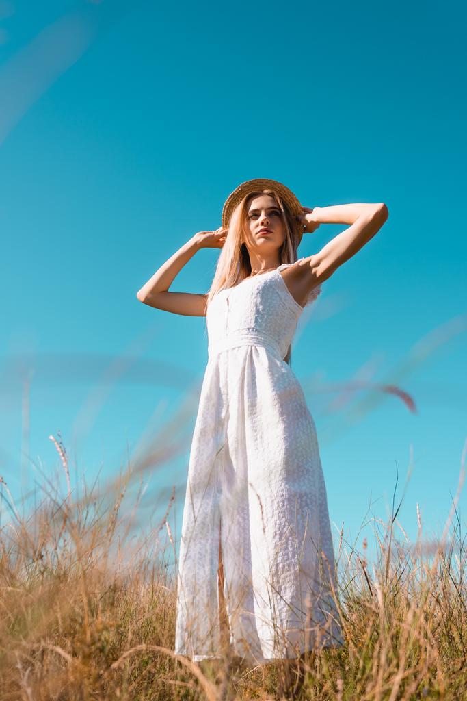 επιλεκτική εστίαση της νεαρής γυναίκας σε λευκό φόρεμα αγγίζοντας ψάθινο καπέλο, ενώ κοιτάζοντας μακριά κατά μπλε ουρανό, άποψη χαμηλή γωνία - Φωτογραφία, εικόνα