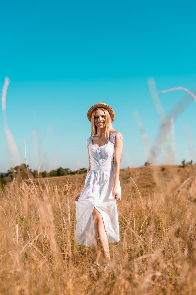 селективное внимание чувственной блондинки в белом платье и соломенной шляпе, смотрящей в камеру, позируя на лугу - Фото, изображение