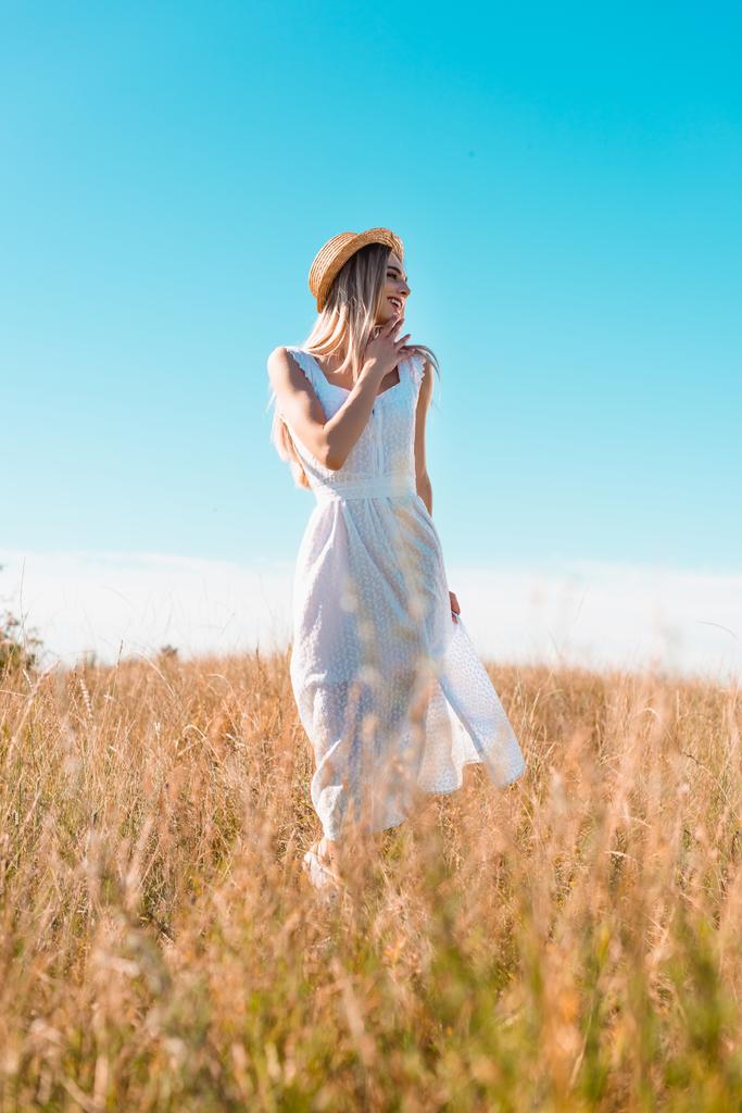 selectieve focus van stijlvolle vrouw in witte jurk en stro hoed aanraken kin en weg te kijken in het veld tegen de blauwe lucht - Foto, afbeelding